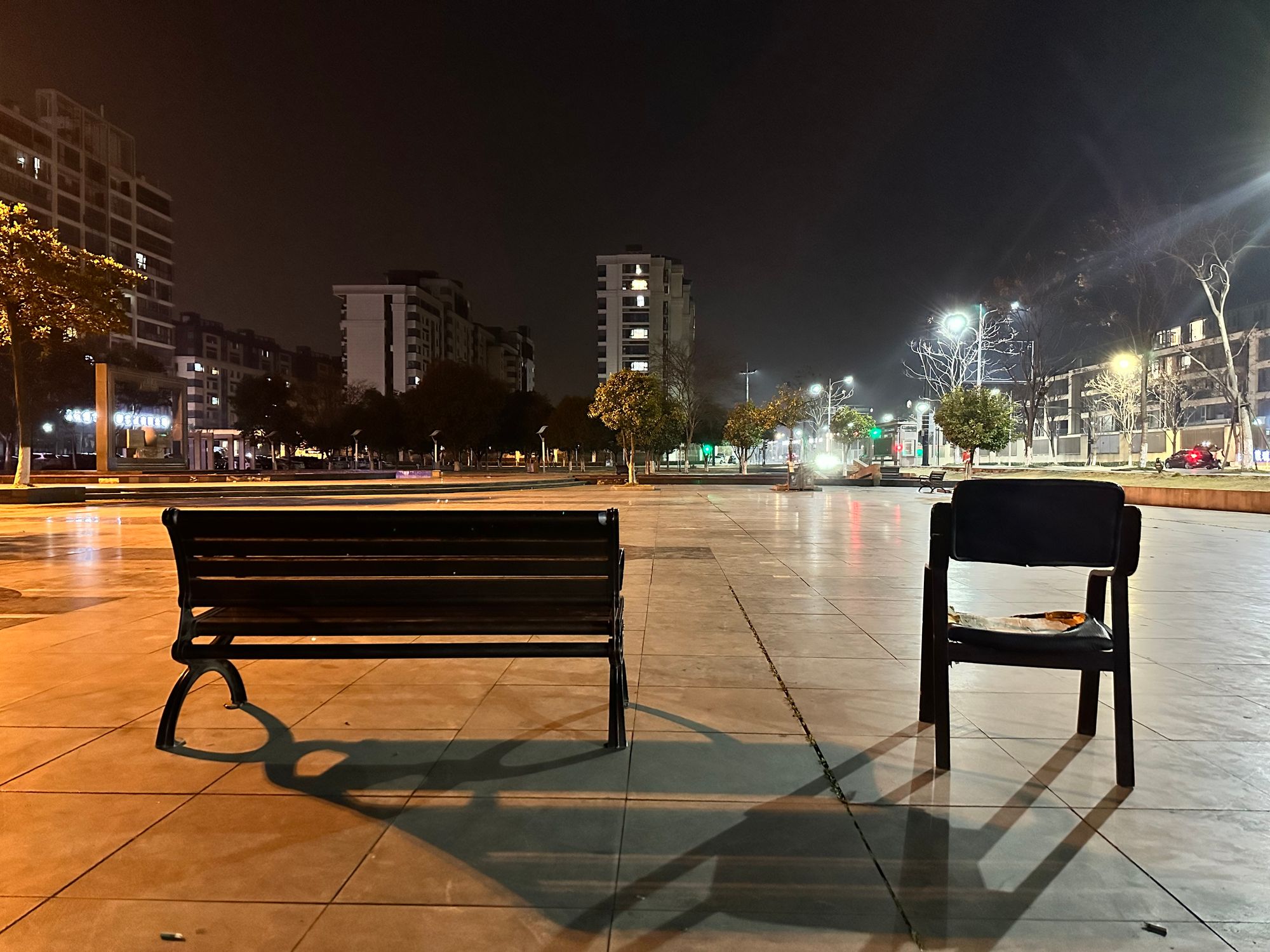 An empty bench and an empty chair next to each other on a square, at night.